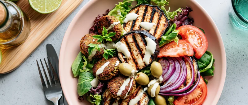 Overhead view of a pink bowl filled with falafel, grilled eggplant, tomatoes, onions, olives and parsley on a gray countertop