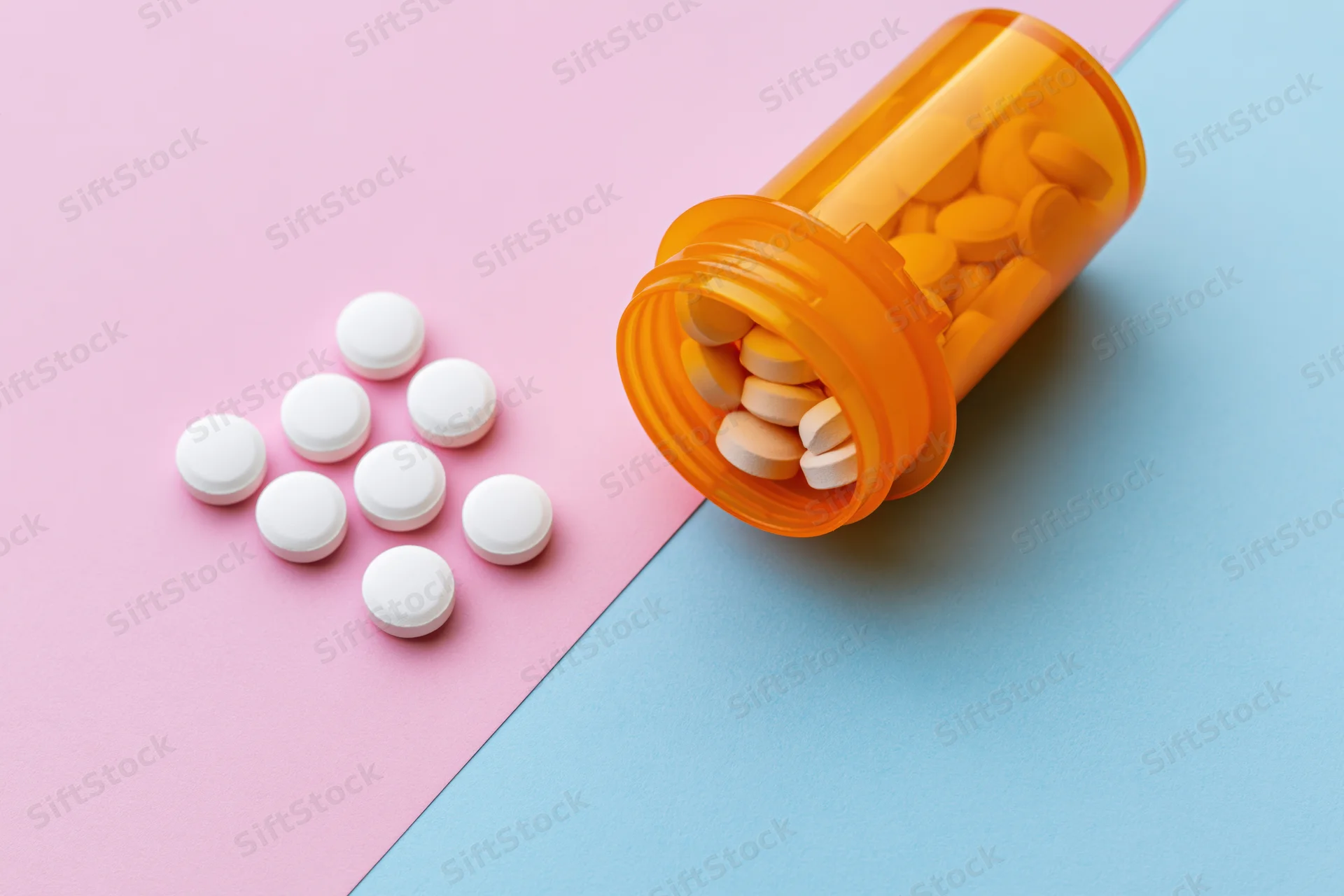 Orange prescription bottle tipped over with white pills spilled onto pink and blue surface