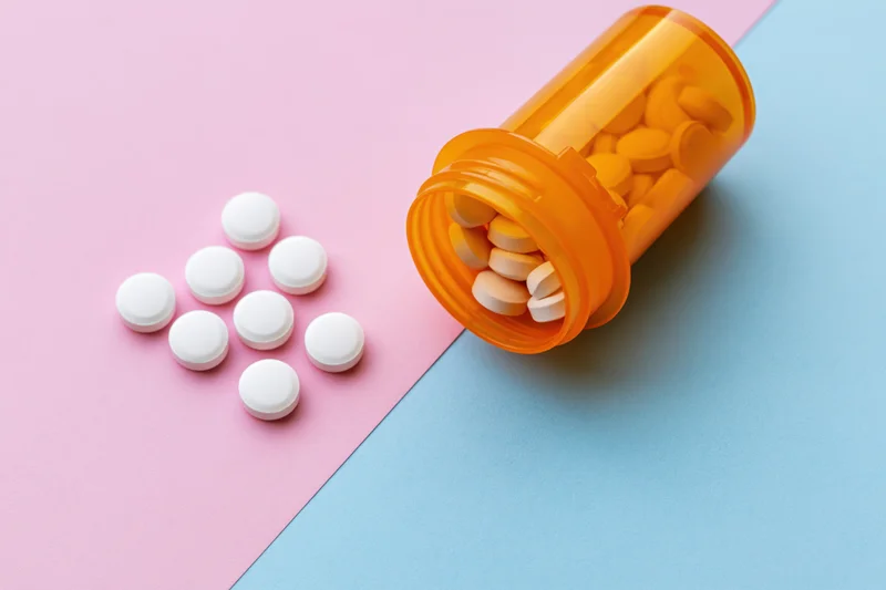 Orange prescription bottle tipped over with white pills spilled onto pink and blue surface
