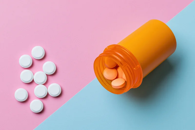 Open orange prescription bottle with scattered white pills on pink and blue surface