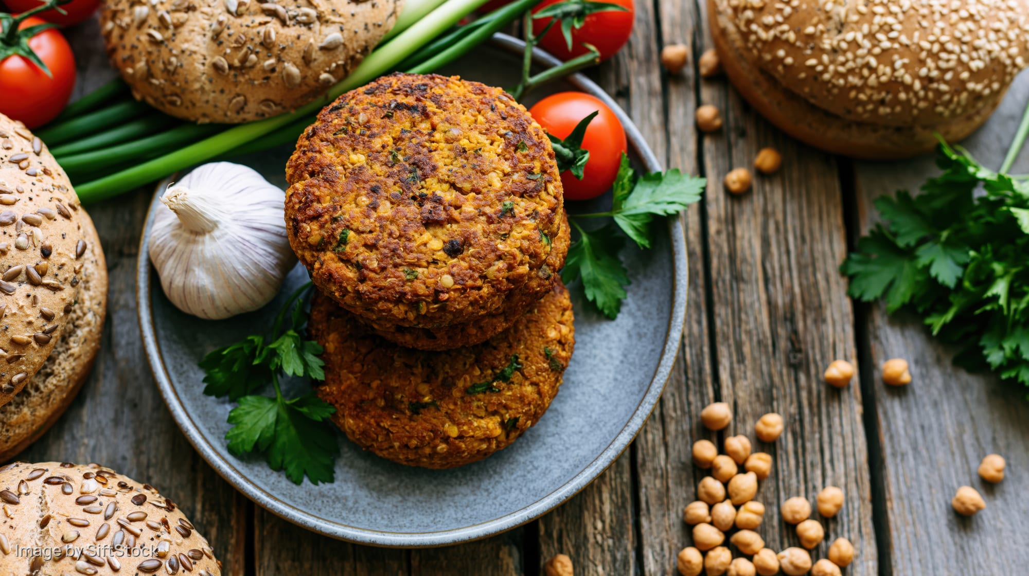 Two stacked homemade chickpea and lentil veggie burgers on a plate surrounded by fresh vegetables and buns on a wooden table