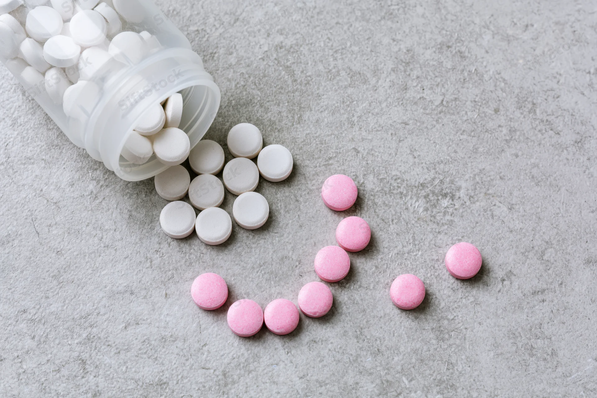 Spilled white and pink tablets from an overturned plastic pill bottle on a gray textured surface