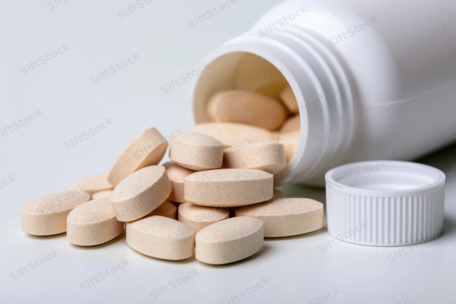 Beige tablets spilling from an overturned white plastic supplement bottle on a plain surface