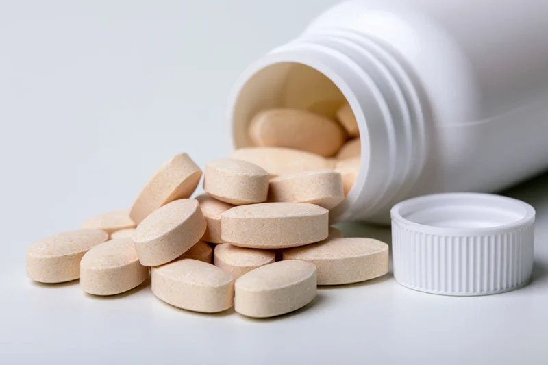 Beige tablets spilling from an overturned white plastic supplement bottle on a plain surface