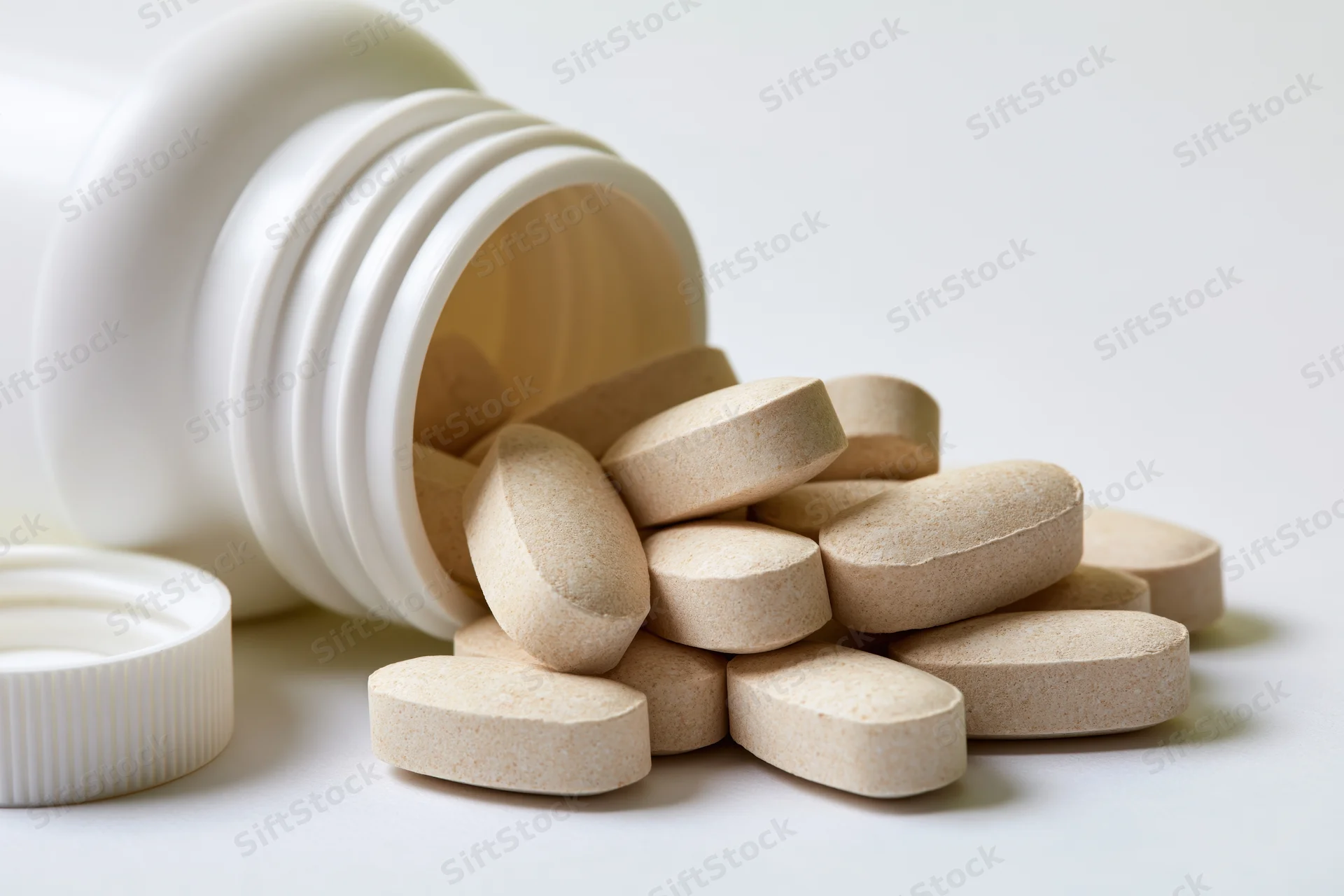 Overturned white plastic supplement bottle with beige tablets spilled onto a plain surface