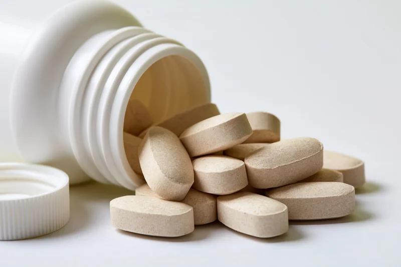 Overturned white plastic supplement bottle with beige tablets spilled onto a plain surface