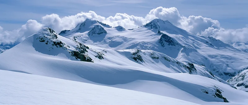 Snow-covered alpine mountains under a partly cloudy sky with rugged peaks and glacier formations