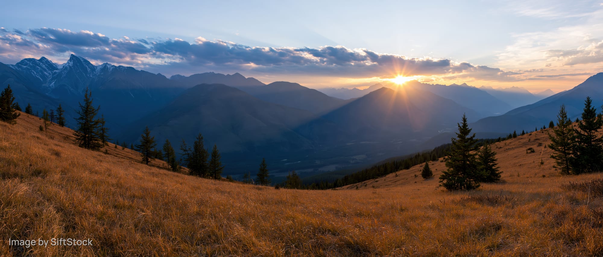 Panoramic alpine landscape at golden hour sunset with layered mountains and grassy meadow in foreground