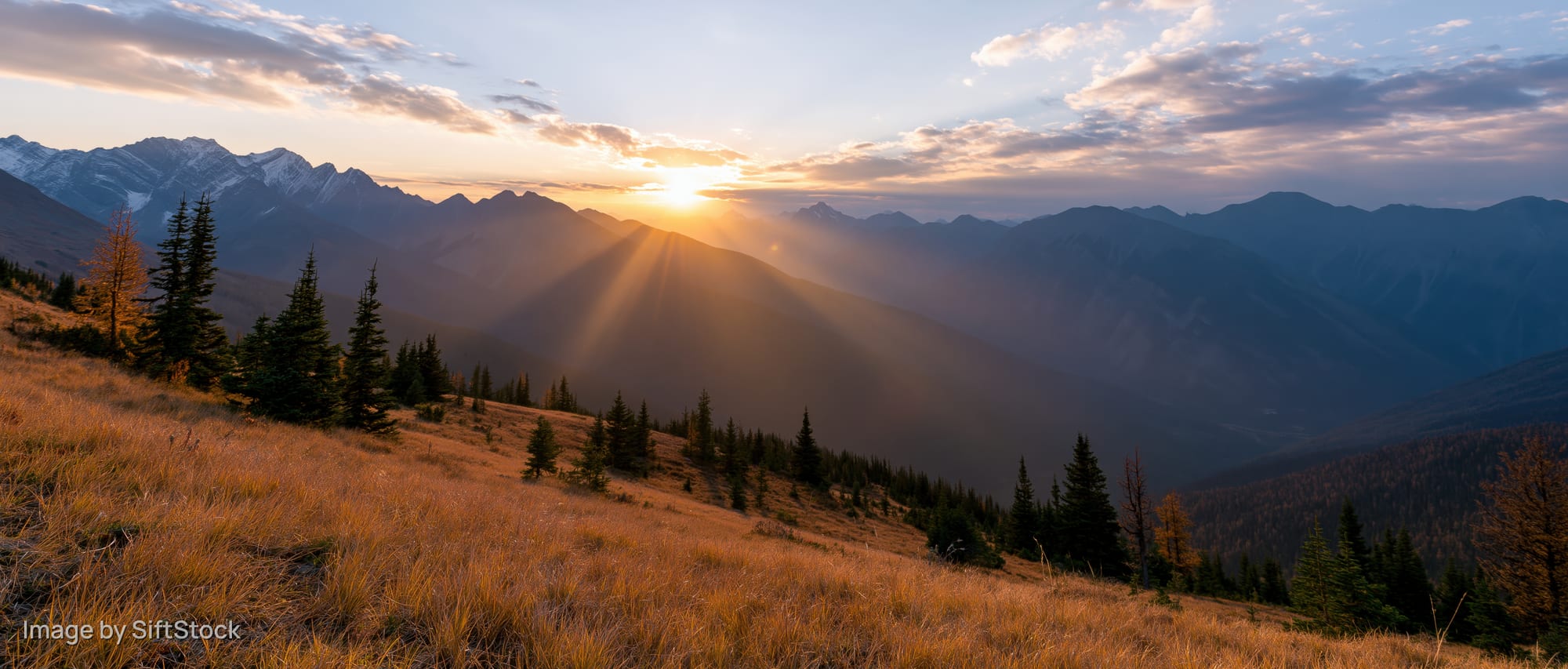 Panoramic alpine landscape at golden hour sunset with sun rays over layered mountains and pine forested valley