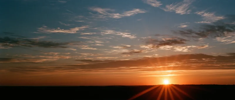 Sunrise over a dark horizon with sunbeams and scattered clouds in a panoramic sky