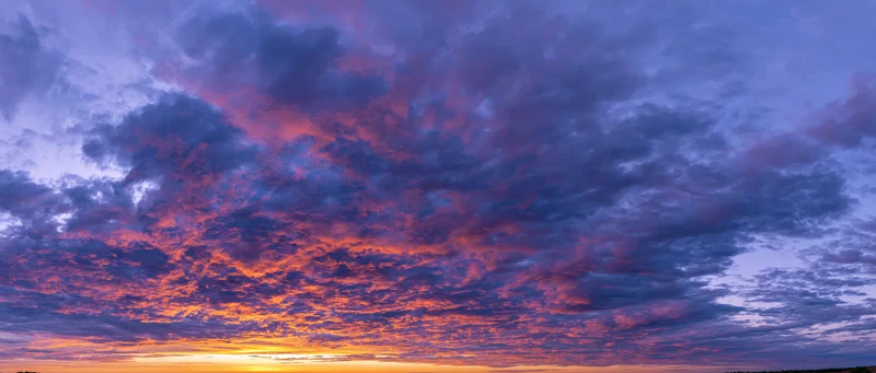 Panoramic sky at sunset with dramatic cumulus clouds and vibrant orange purple hues above the horizon