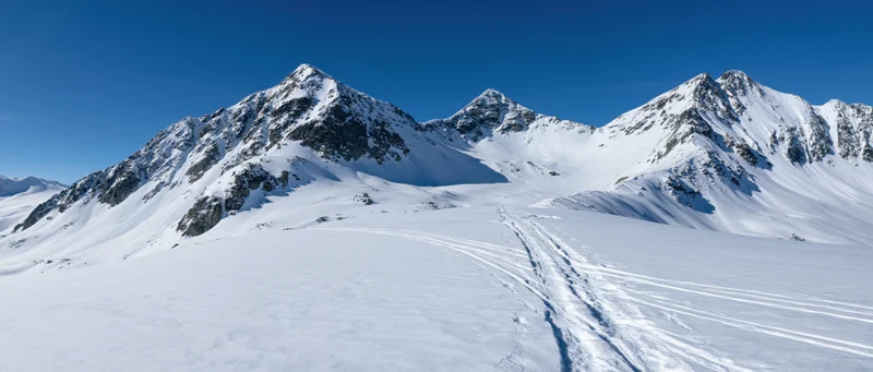 Snow-covered alpine peaks rising above a wide valley with ski tracks under clear blue sky