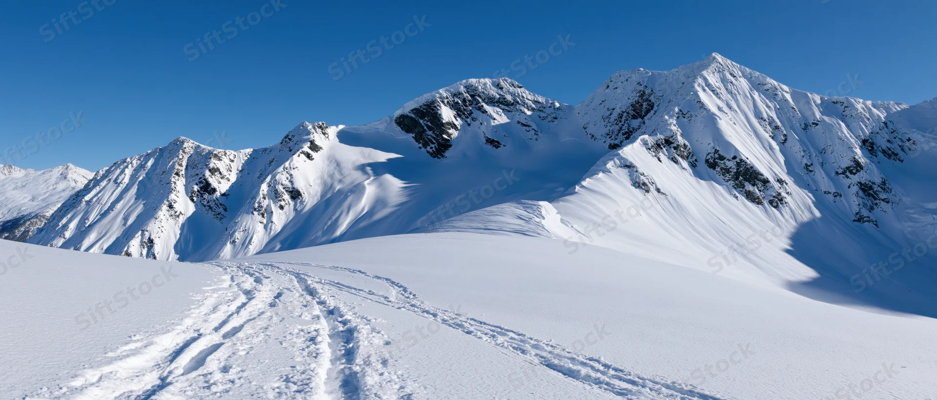 Snow-covered alpine peaks rising above a snowy valley with ski tracks leading toward them under a clear blue sky