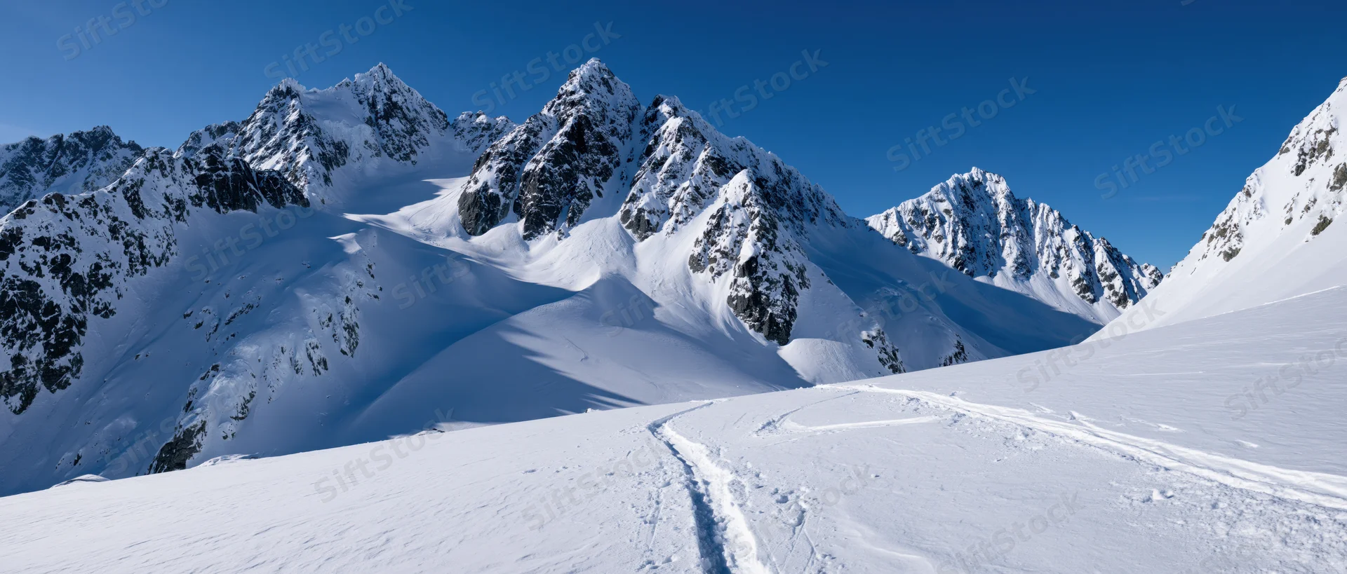 Snow-covered alpine peaks rising above a snowy valley with ski tracks leading toward the ridges under a clear blue sky