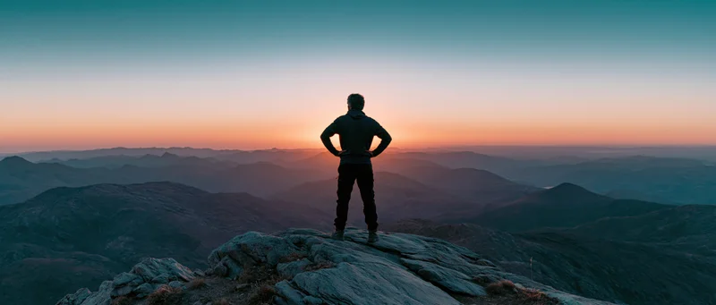 Man standing on rocky mountain summit with hands on hips overlooking layered valleys at sunset