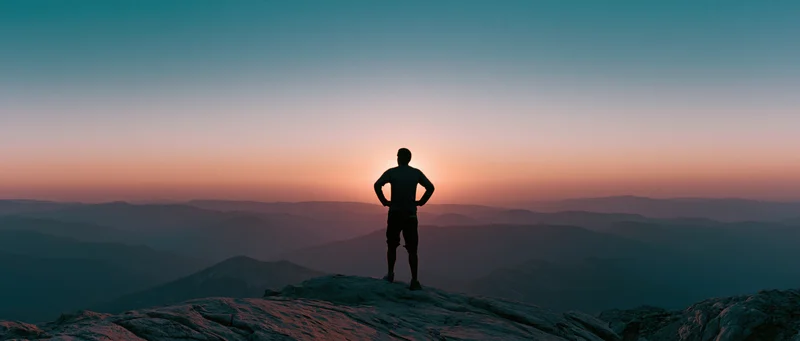 Man standing alone on rocky mountain summit at sunset with hands on hips