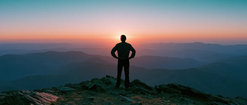 Man standing alone on rocky mountain summit at sunset with hands on hips overlooking layered valleys