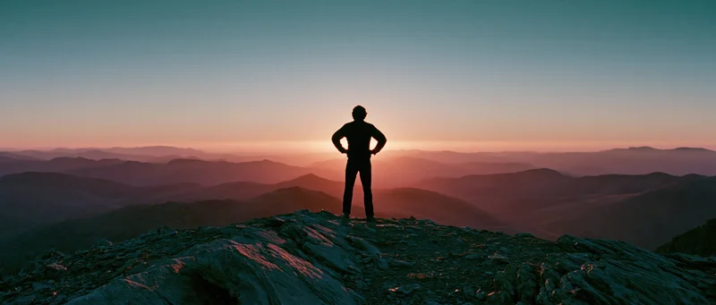 Silhouette of a person standing alone on a rocky mountain summit at sunset with hands on hips overlooking layered valleys