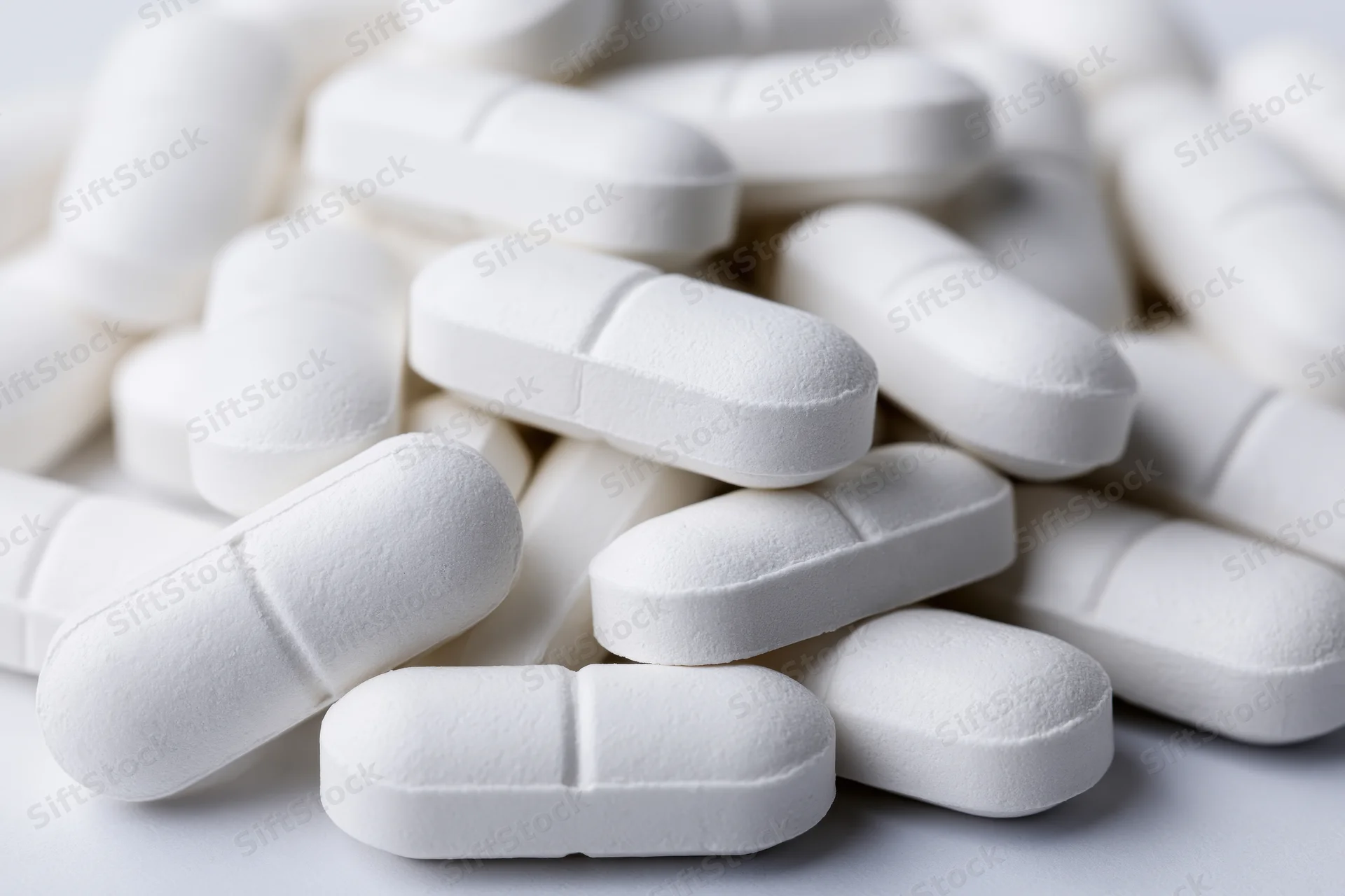Pile of white oval pharmaceutical tablets scattered loosely on a plain surface