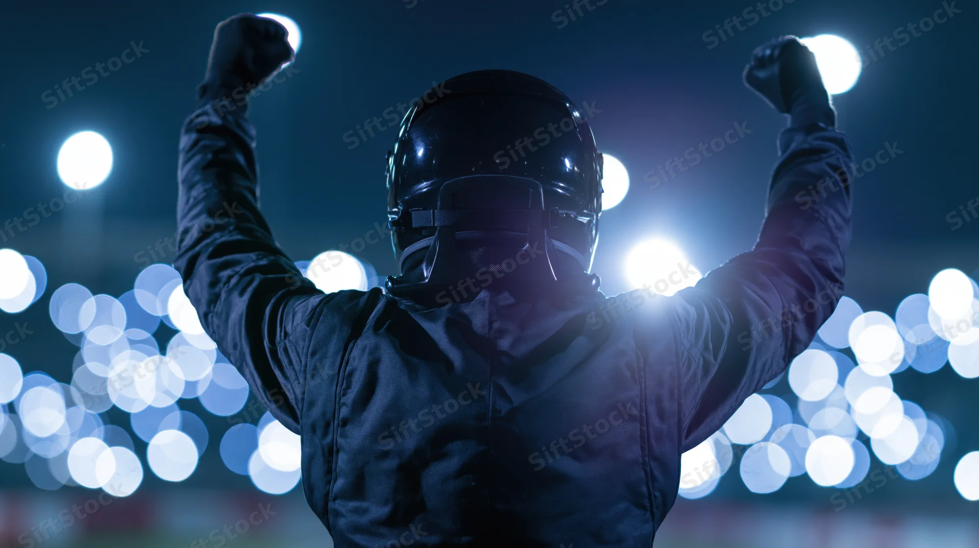 Racing driver in black suit and helmet raising arms in victory at night stadium