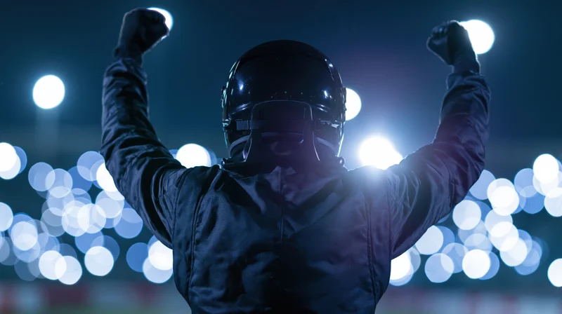 Racing driver in black suit and helmet raising arms in victory at night stadium