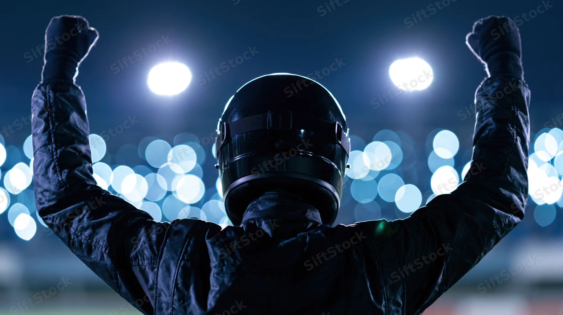Racing driver in black suit and helmet raising arms in victory at night stadium