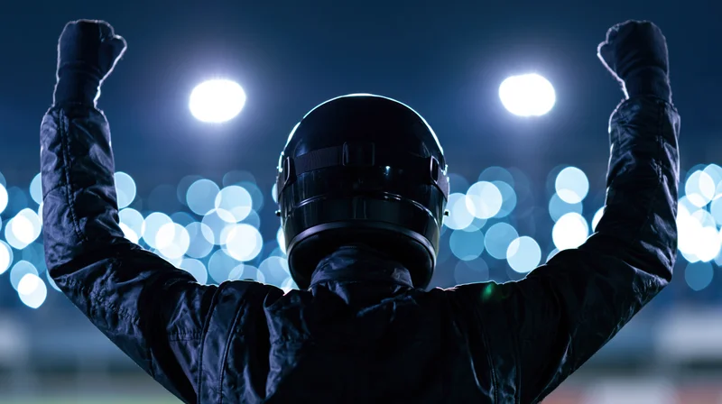 Racing driver in black suit and helmet raising arms in victory at night stadium