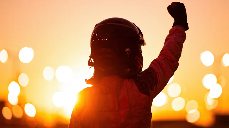Racing driver in full helmet and firesuit raising fist in celebration at sunset
