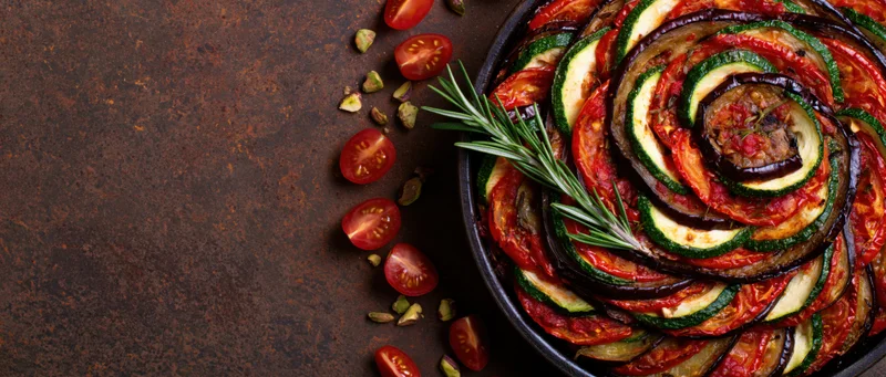Ratatouille arranged in a spiral pattern in a round baking dish with cherry tomatoes and rosemary on a dark textured surface