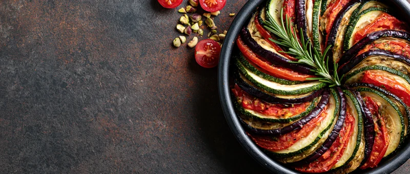 Ratatouille in a round baking dish with layered vegetables and rosemary garnish on a dark rustic surface