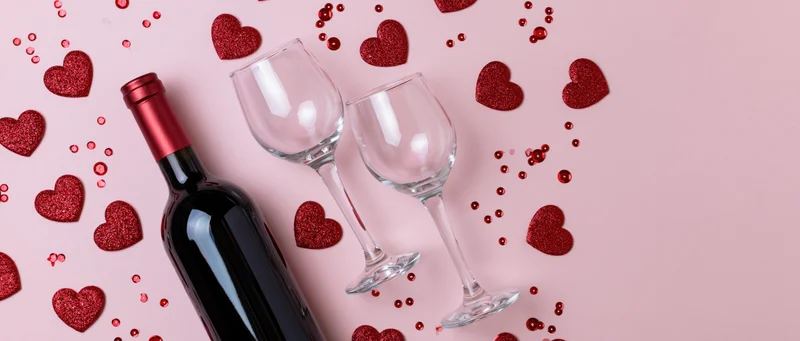 Red wine bottle and two empty tilted glasses on pink surface with glitter hearts scattered around