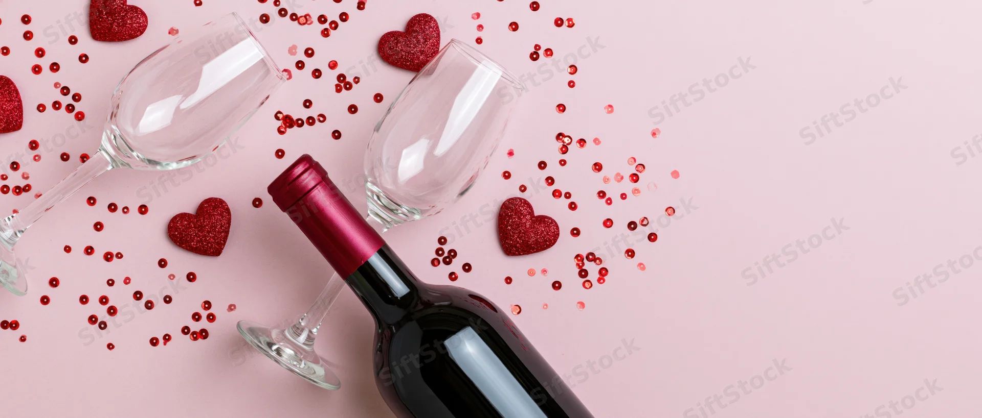 Red wine bottle lying on its side next to two empty tilted wine glasses on pink surface with glitter hearts and confetti