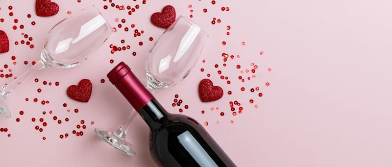 Red wine bottle lying on its side next to two empty tilted wine glasses on pink surface with glitter hearts and confetti