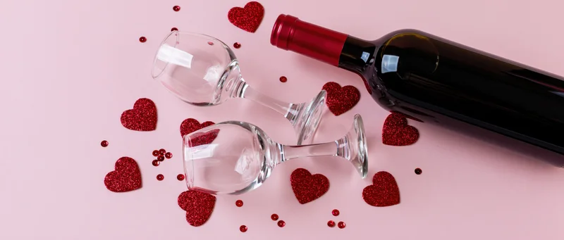 Red wine bottle and two empty glasses with glitter hearts on pink surface