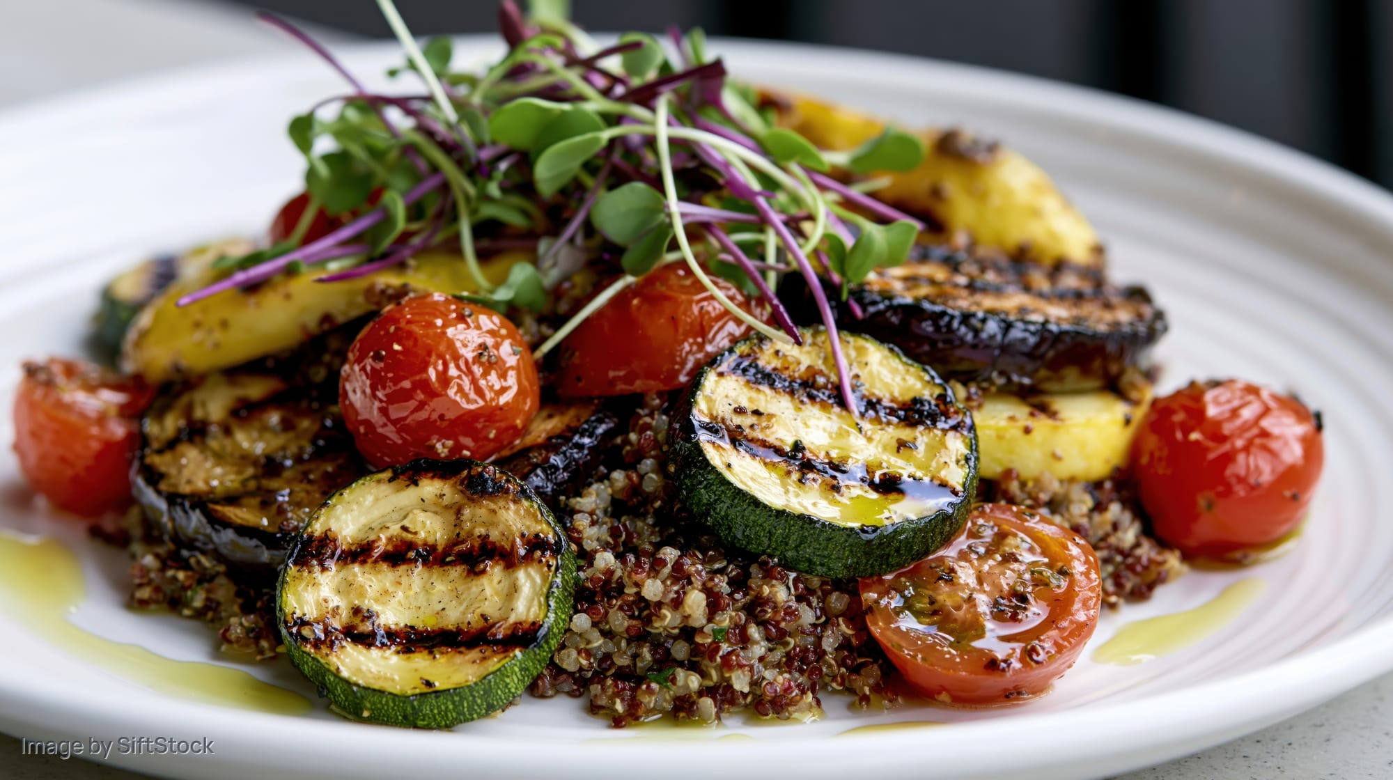 Grilled vegetables and quinoa salad served on a white plate with microgreens and olive oil drizzle