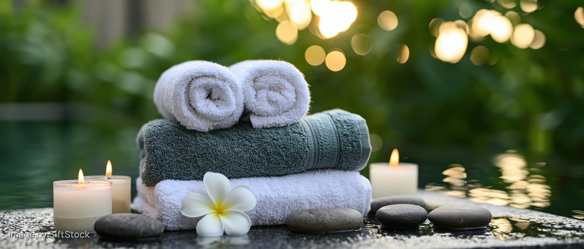 Stacked spa towels with candles and stones beside a pool at evening