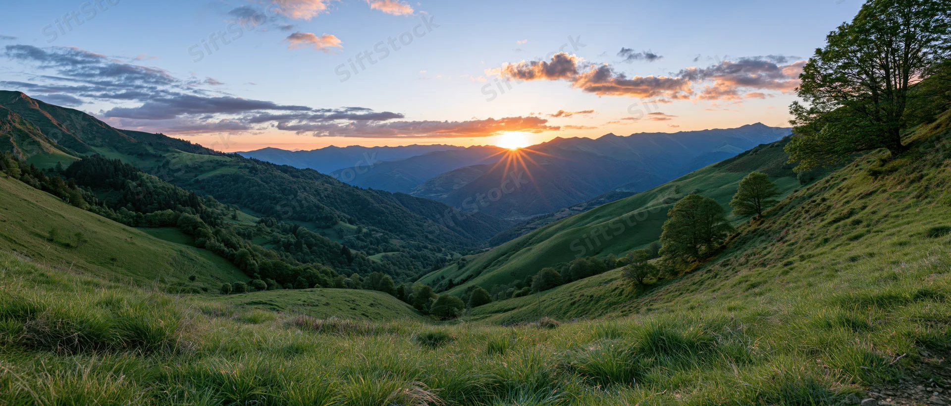 Wide view of rolling green hills and mountain valley at sunset with sun low on horizon