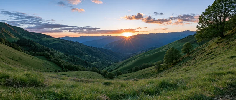 Wide view of rolling green hills and mountain valley at sunset with sun low on horizon