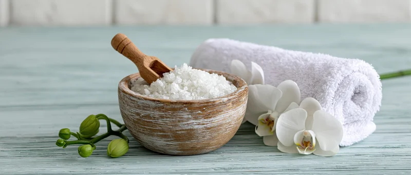 Coarse bath salts in a rustic wooden bowl with white orchid and towel on a light blue wooden surface
