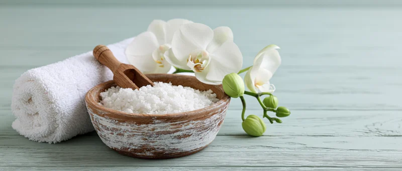 White orchid and bath salts in a rustic wooden bowl beside a towel on a painted wood surface