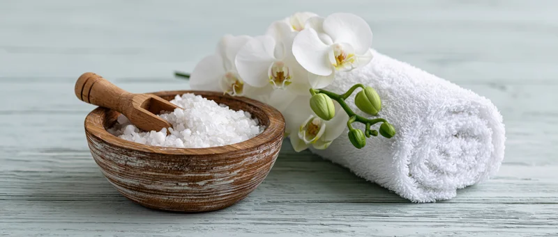 Wooden bowl with bath salts and orchid beside rolled towel on light blue wooden surface
