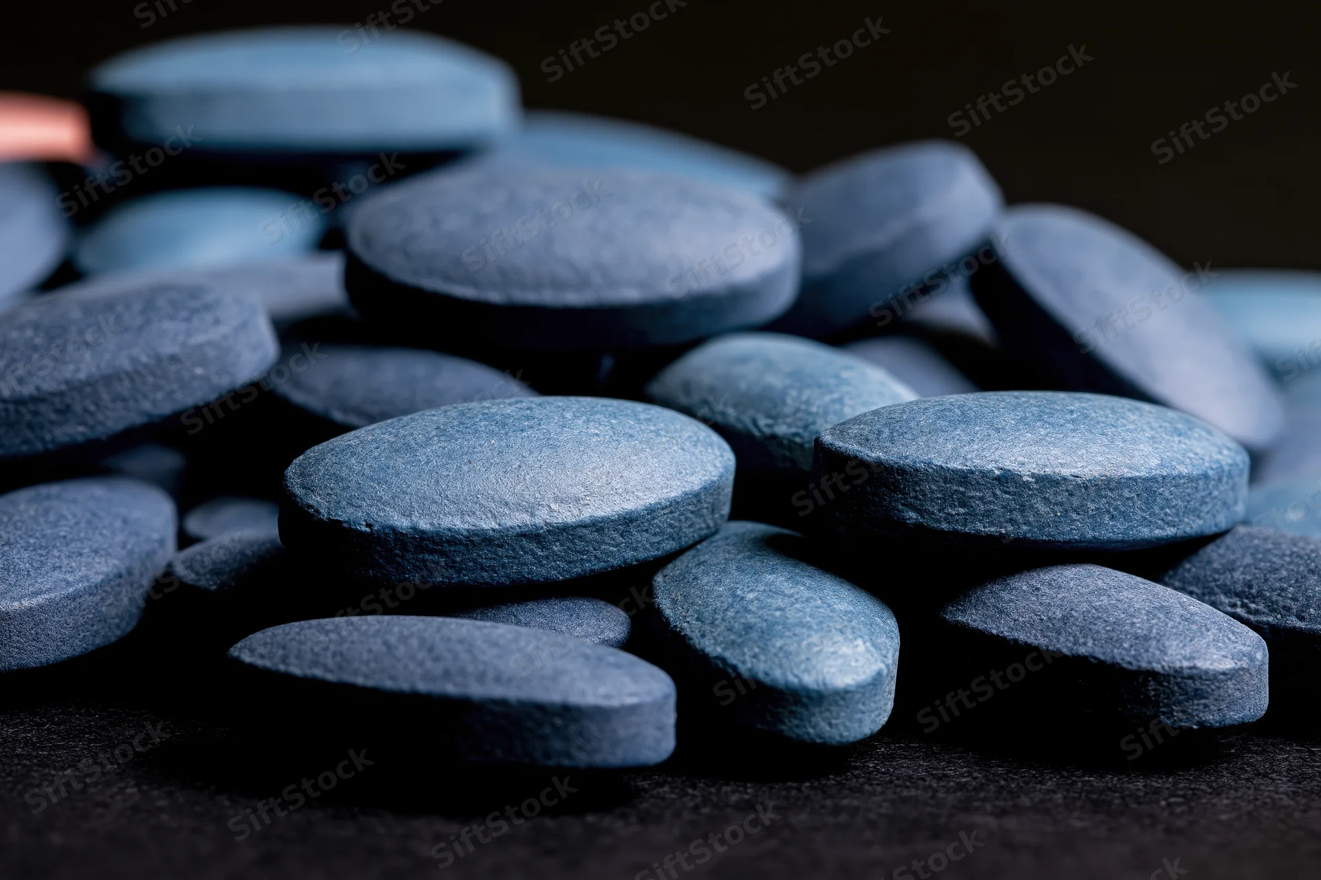 Scattered oval blue pharmaceutical tablets on dark surface