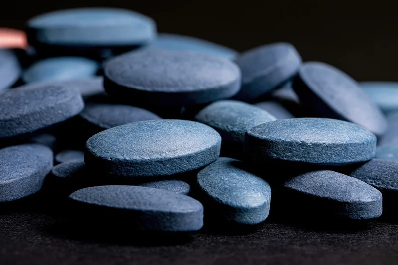 Scattered oval blue pharmaceutical tablets on dark surface