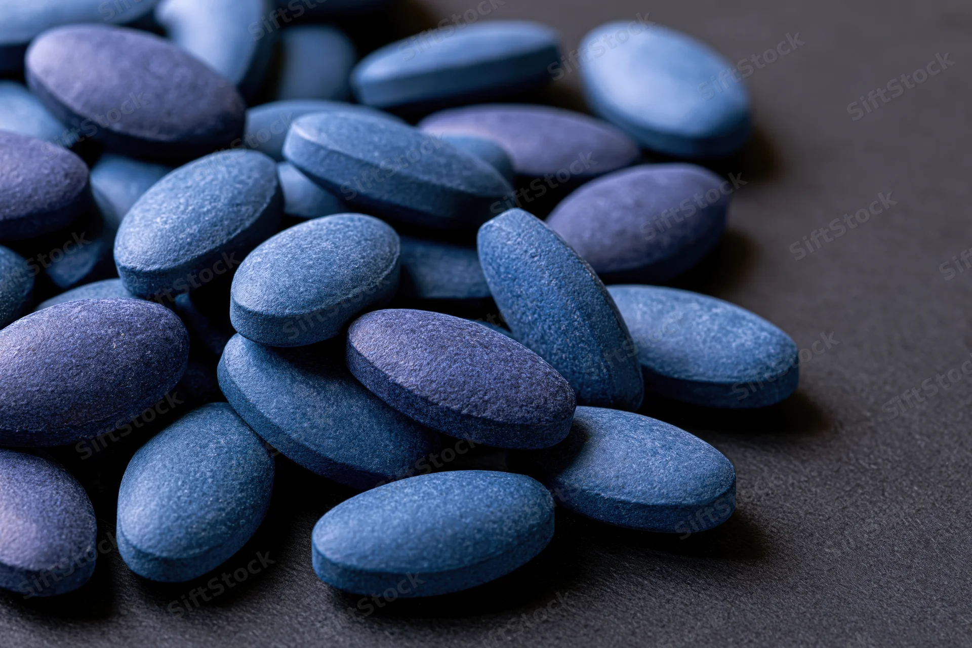 Scattered oval blue pharmaceutical tablets on dark surface