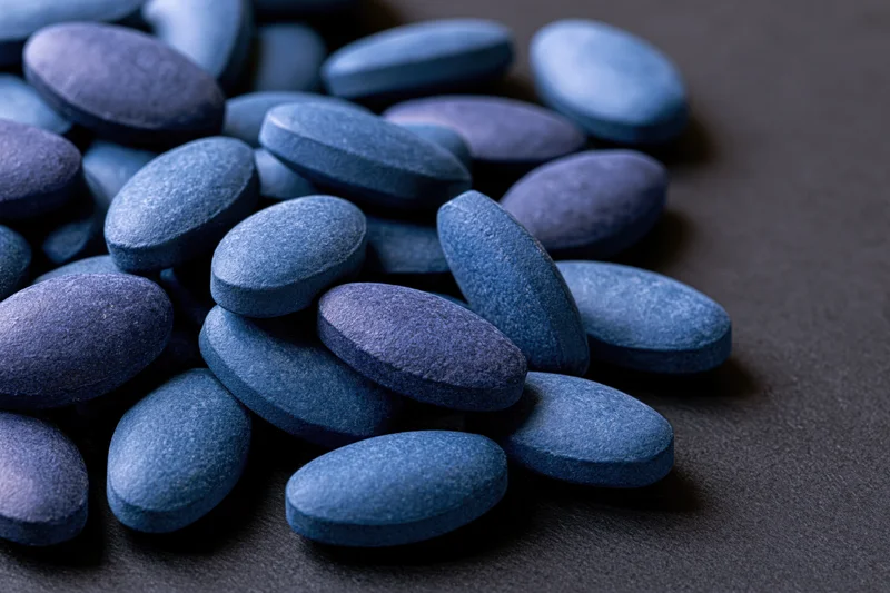 Scattered oval blue pharmaceutical tablets on dark surface