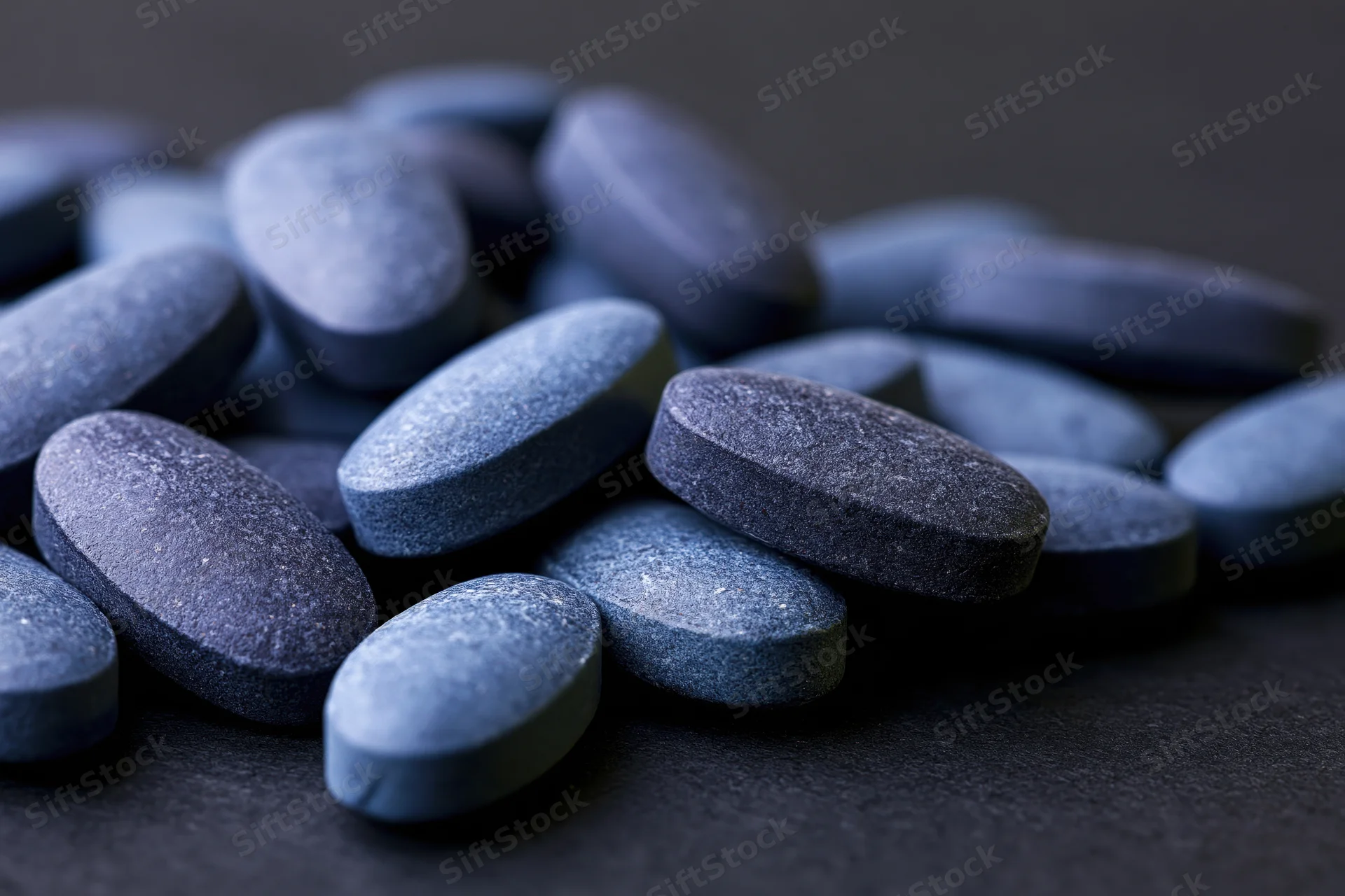 Scattered oval blue pharmaceutical tablets on dark surface