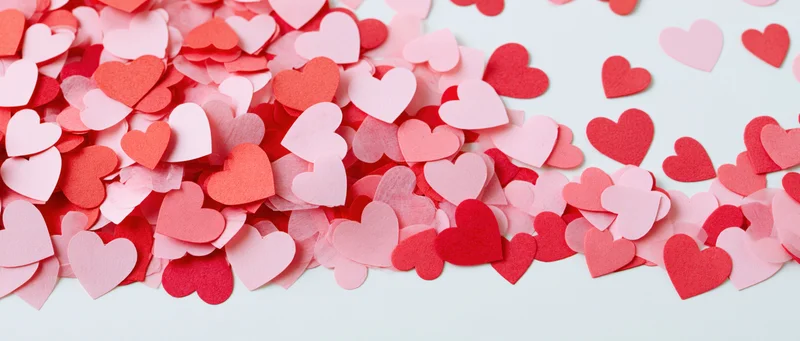 Scattered paper heart confetti in red and pink tones on a light background with copyspace