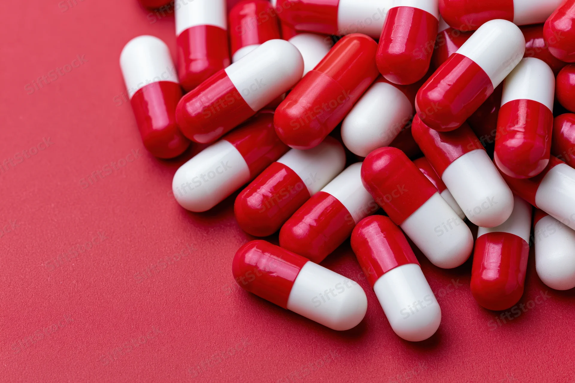 Scattered red and white two-tone capsule pills piled on a red surface