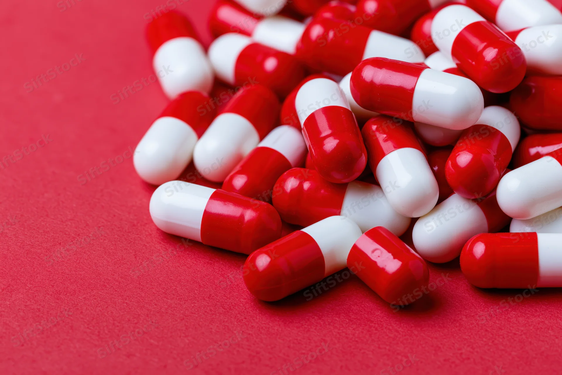 Scattered red and white two-tone capsule pills piled on a red surface