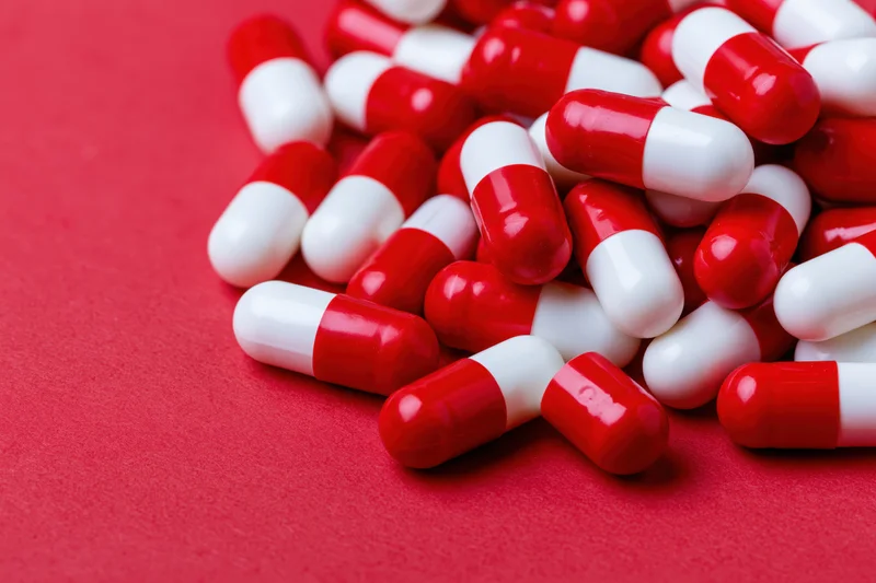 Scattered red and white two-tone capsule pills piled on a red surface